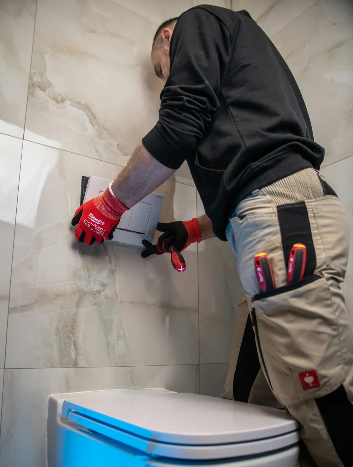 Plombier en gants rouges posant la plaque de commande d'un WC suspendu sur un mur carrelé beige