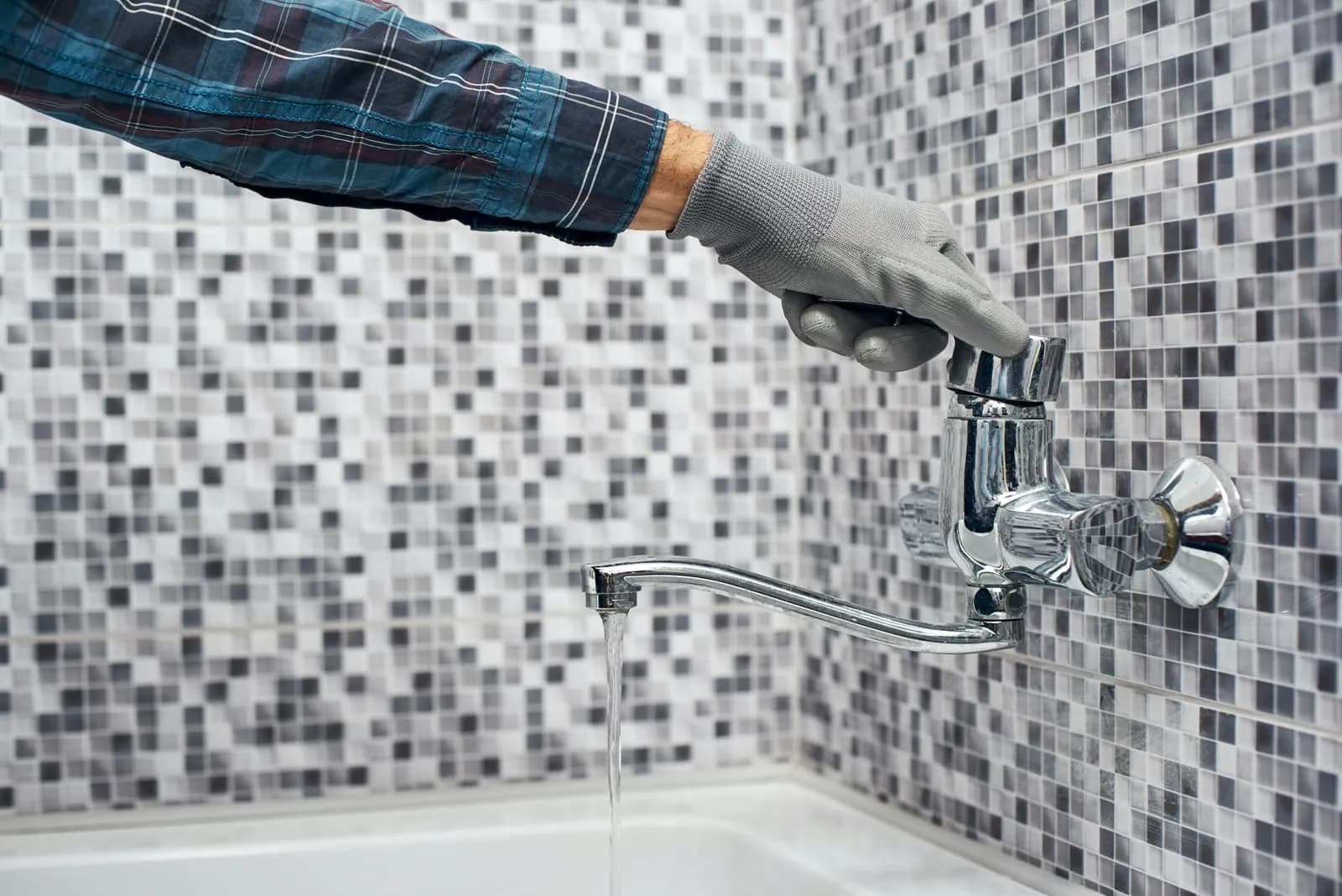 Main gantée de gris vérifiant un mitigeur chromé qui laisse couler un filet d'eau sur un mur de mosaïque