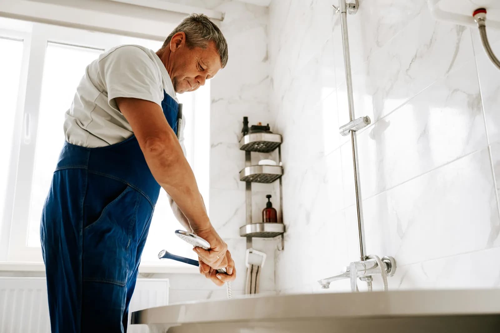 Homme en salopette bleue serrant un raccord de flexible de douche au-dessus d'une baignoire blanche
